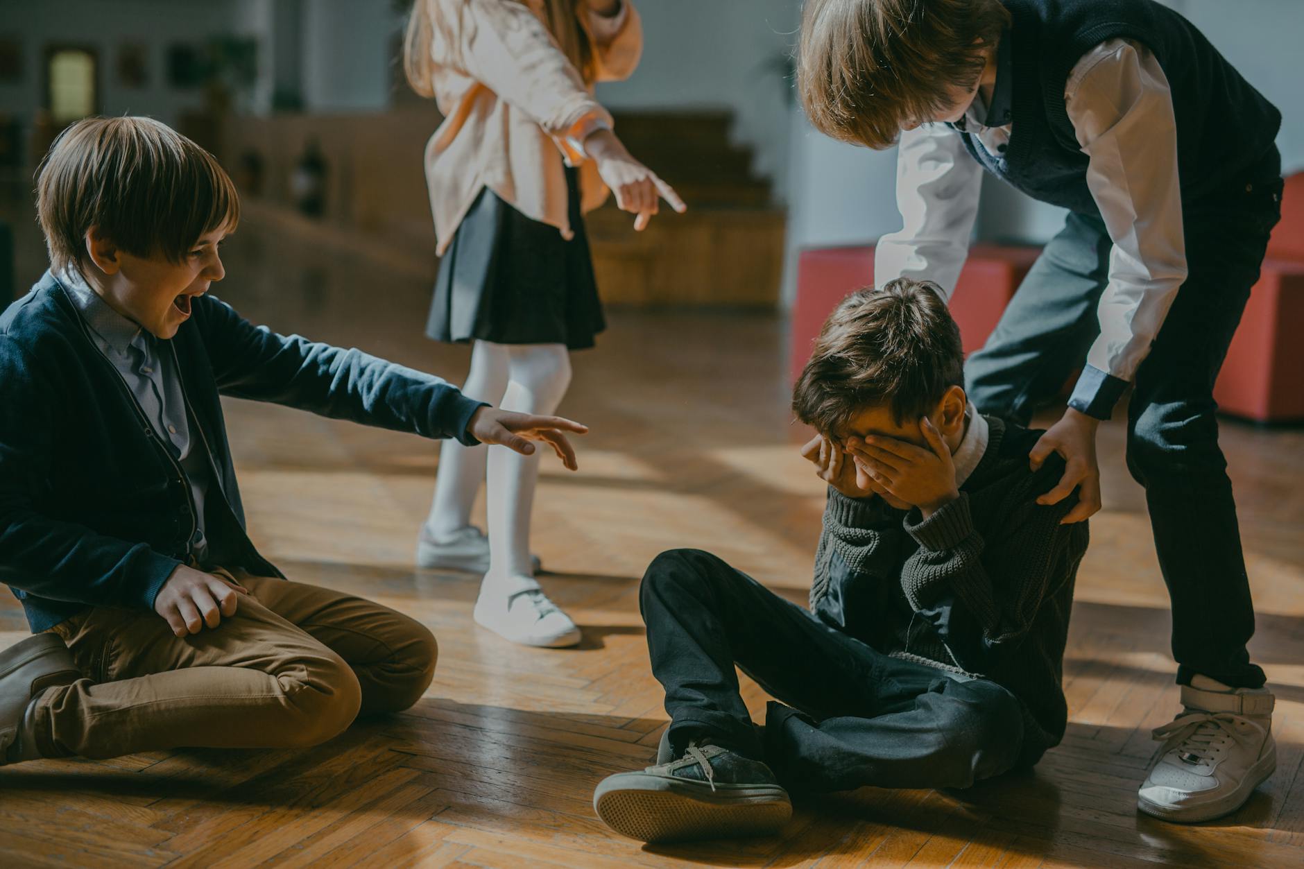 children finger pointing at a boy sitting on a wooden floor